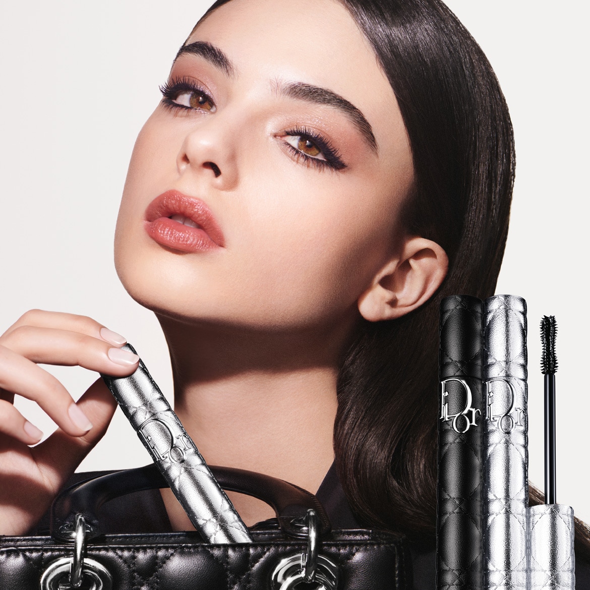 A képen egy nő látható, aki Dior sminktermékeket használ, a szempillaspirál ezüst és fekete színű csomagolásban kapható, a Dior logóval ellátva.