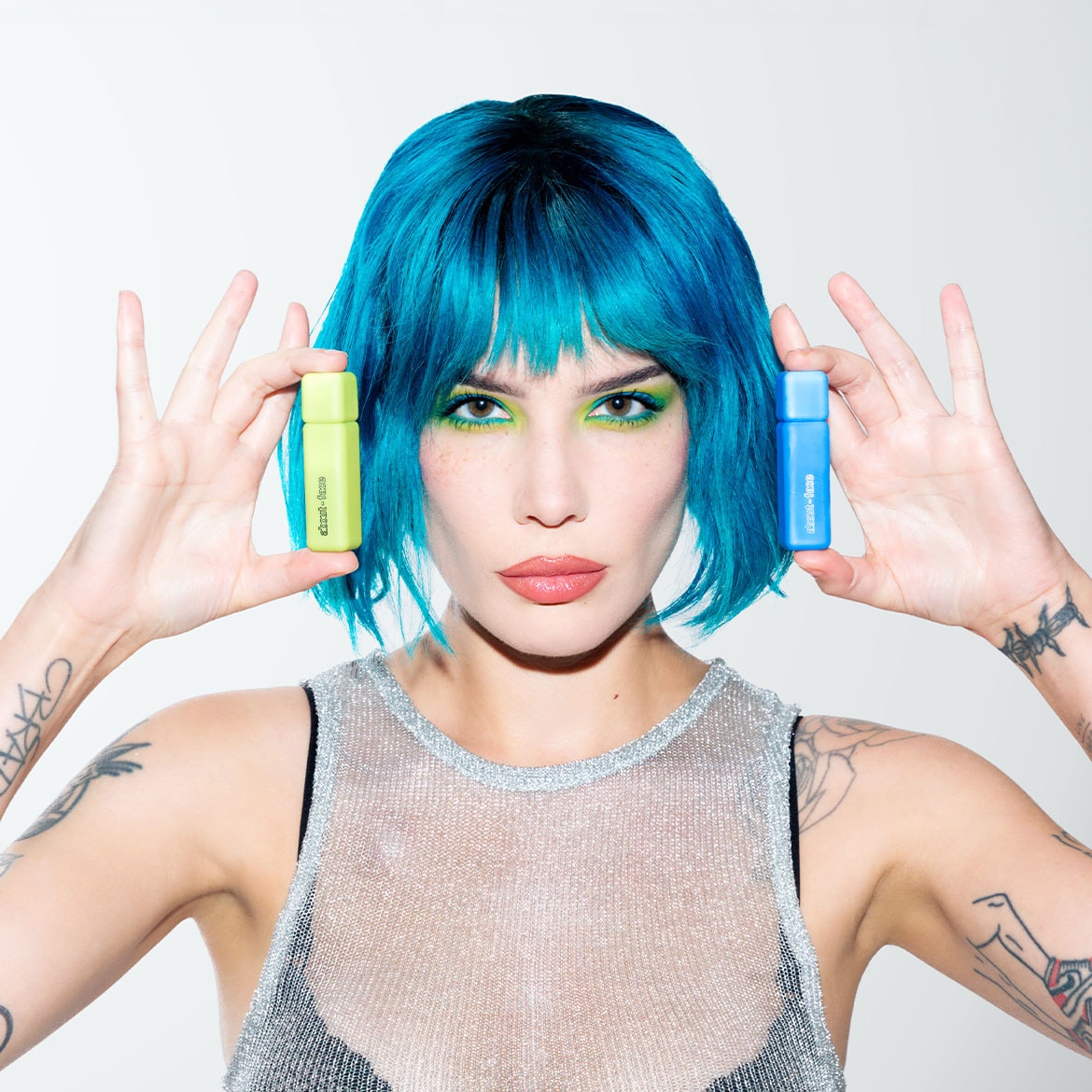 A kék hajú, tetovált nő ezüst színű felsőben zöld és kék színű easy-to-face termékeket tart a kezében.