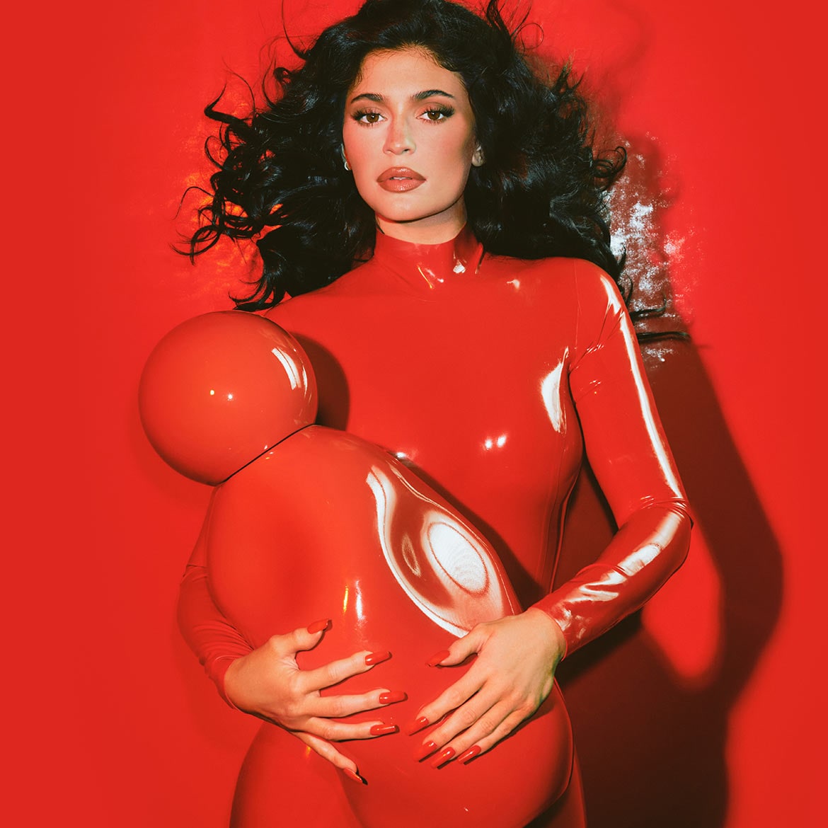 Kylie Jenner vörös, fényes latex ruhában, kezében egy piros, gömb alakú tárgyat tartva, a háttér is vörös.