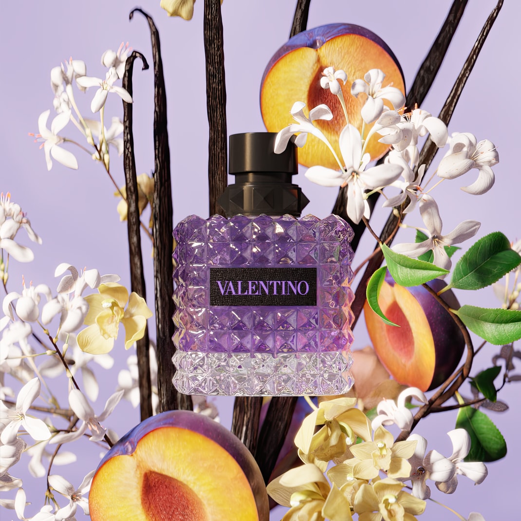 A Valentino Donna Born In Roma lila színű, sokszögű üvegben, fekete kupakkal, virágok, szilva és vaníliarudak között lebegve.