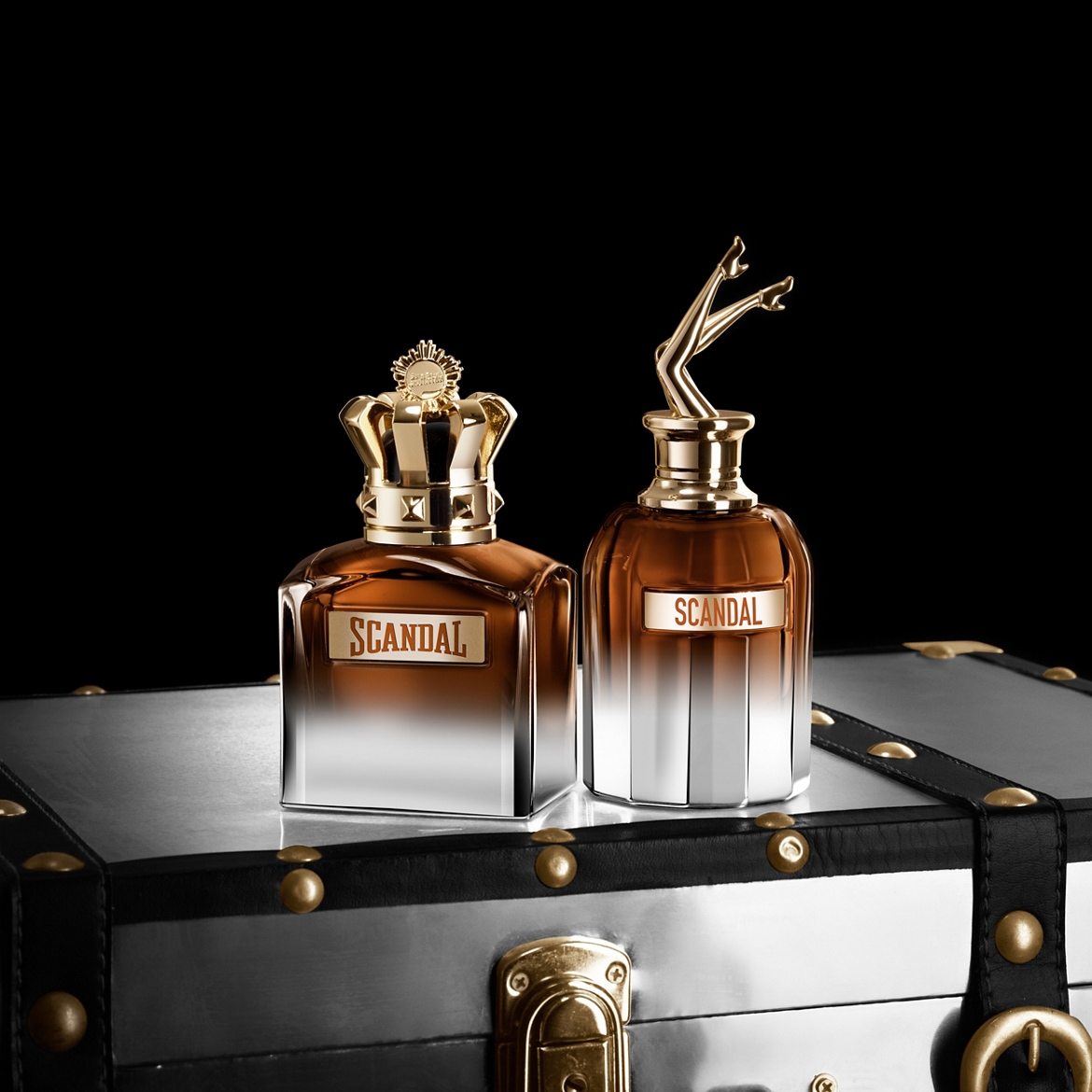 Két Scandal parfümös üveg látható egy ezüstszínű, fekete szegélyű bőröndön, az egyik üveg tetején egy arany színű korona, a másikon pedig egy női láb szobrocskája található.