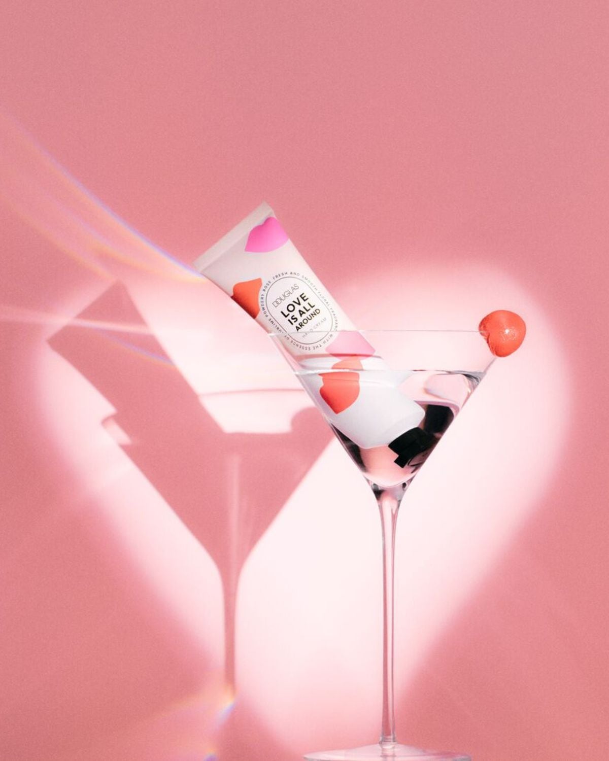 Douglas Love Is All Around kézkrém rózsaszín háttér előtt, martinis pohárban, rózsaszín és narancssárga szív alakú díszítéssel.
