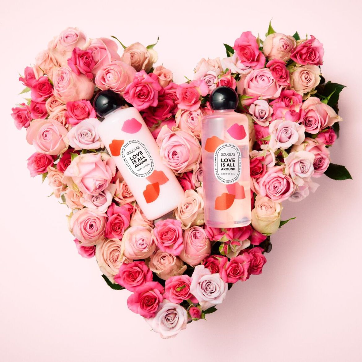 Two Douglas 'Love is All Around' products, a white body lotion and a transparent shower gel, sit nestled in a heart-shaped arrangement of pink and peach roses.