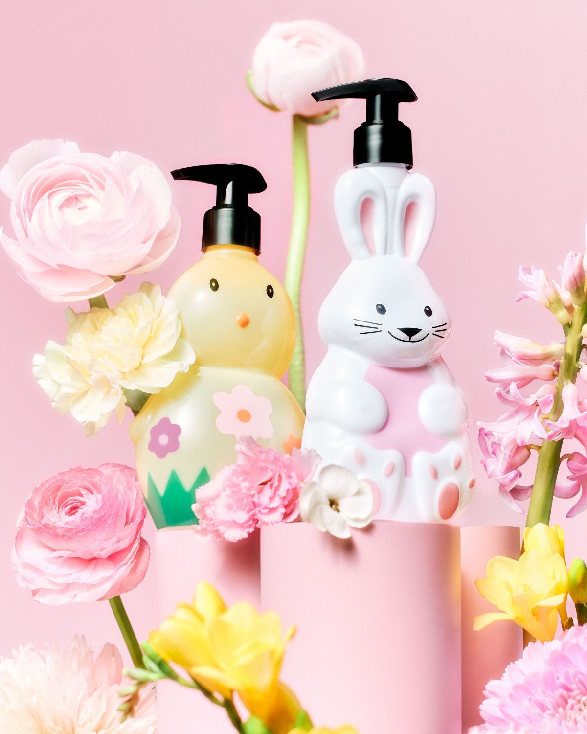 A rózsaszín háttér előtt egy sárga csibe és egy fehér nyuszi alakú szappanadagoló áll, mindkettő fekete pumpával, virágokkal körülvéve.