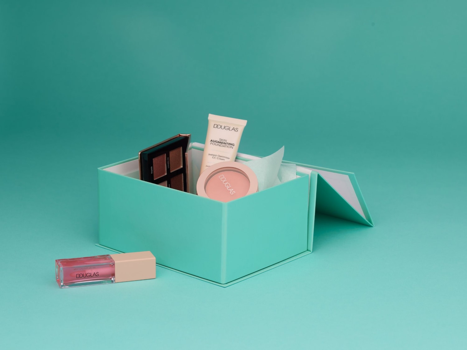 A Douglas márkájú sminktermékekből álló kollekciót tartalmazó, türkizkék dobozban egy szemhéjpúder paletta, egy alapozó, egy pirosító és egy szájfény található.