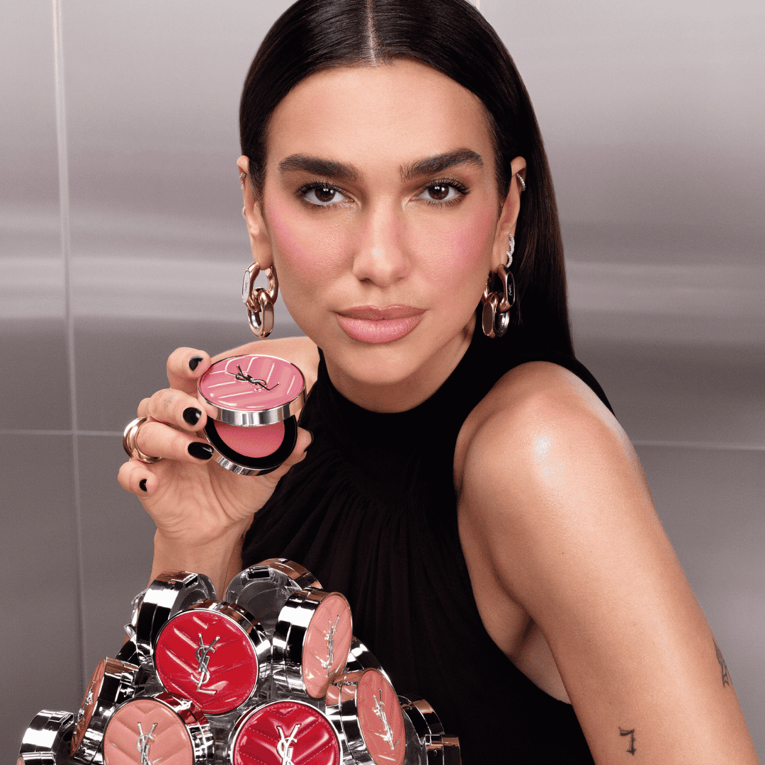 Egy nő tartja a kezében a YSL Beauty pirosító kompaktot, amelynek rózsaszín fedelén a márka logója látható, miközben a háttérben több pirosító kompakt is látható.