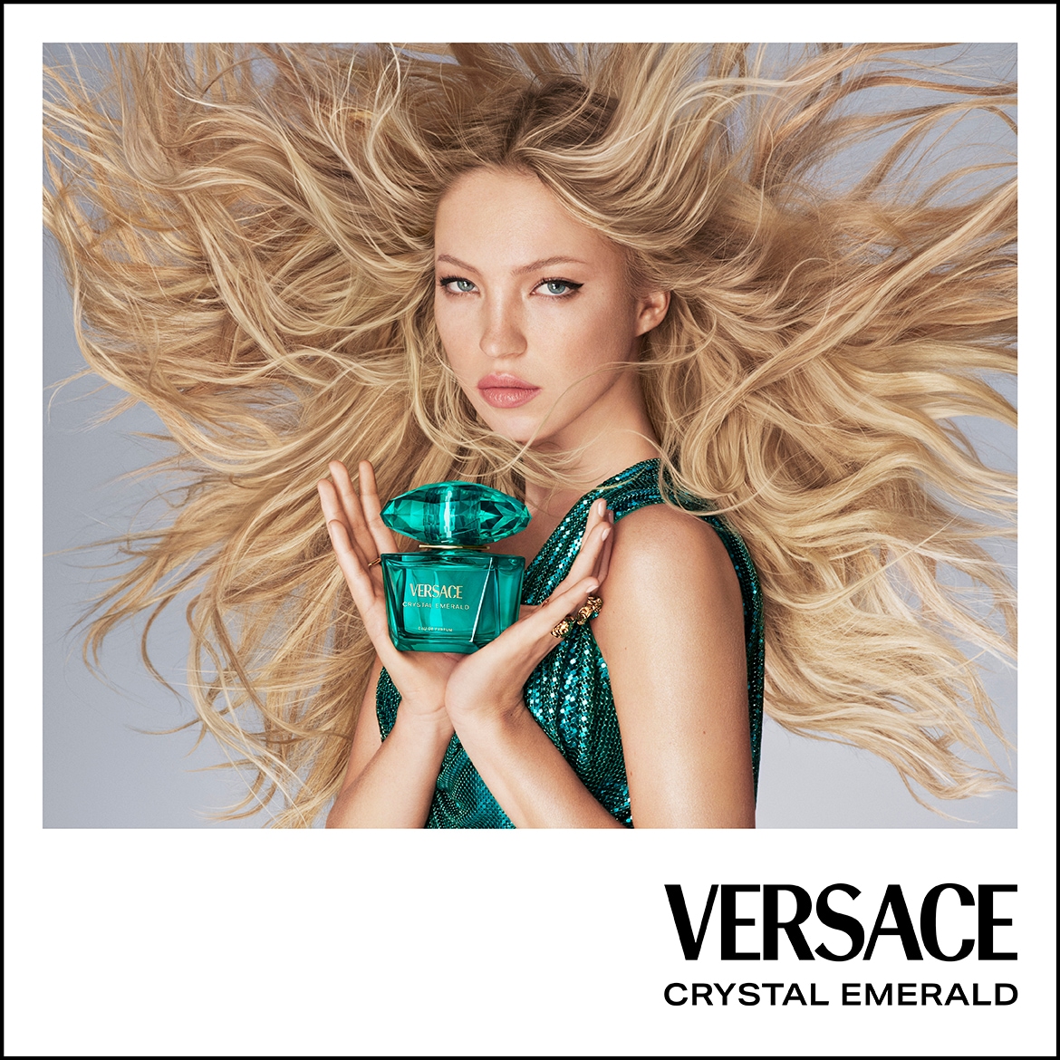 Egy szőke hajú nő csillogó smaragdzöld ruhában tartja a Versace Crystal Emerald parfümöt, melynek üvege egy csiszolt smaragdra emlékeztet.